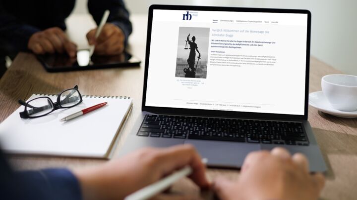 Laptop mit geöffneter Website auf einem Tisch neben Notizblock, Brille und Tasse