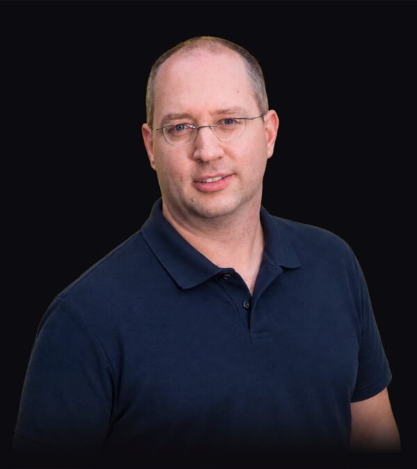 Ein Mann in einem dunkelblauen Poloshirt schaut vor schwarzem Hintergrund in die Kamera