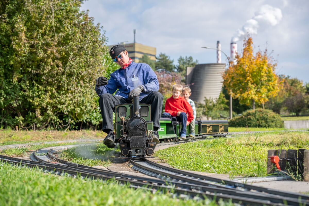 Eine Person fährt eine kleine Bahn mit sitzenden Kindern über ein Gleis im Freien
